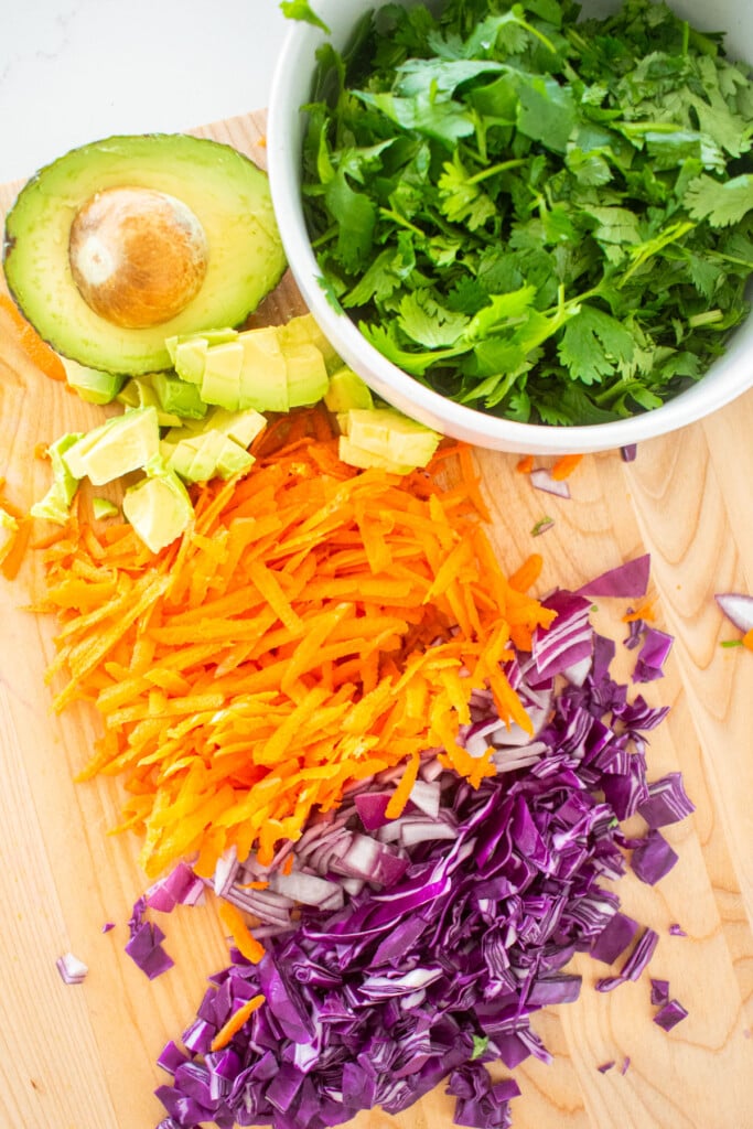 chopped cilantro, purple cabbage, avocado red onion, grated carrots, on a wood cutting board.
