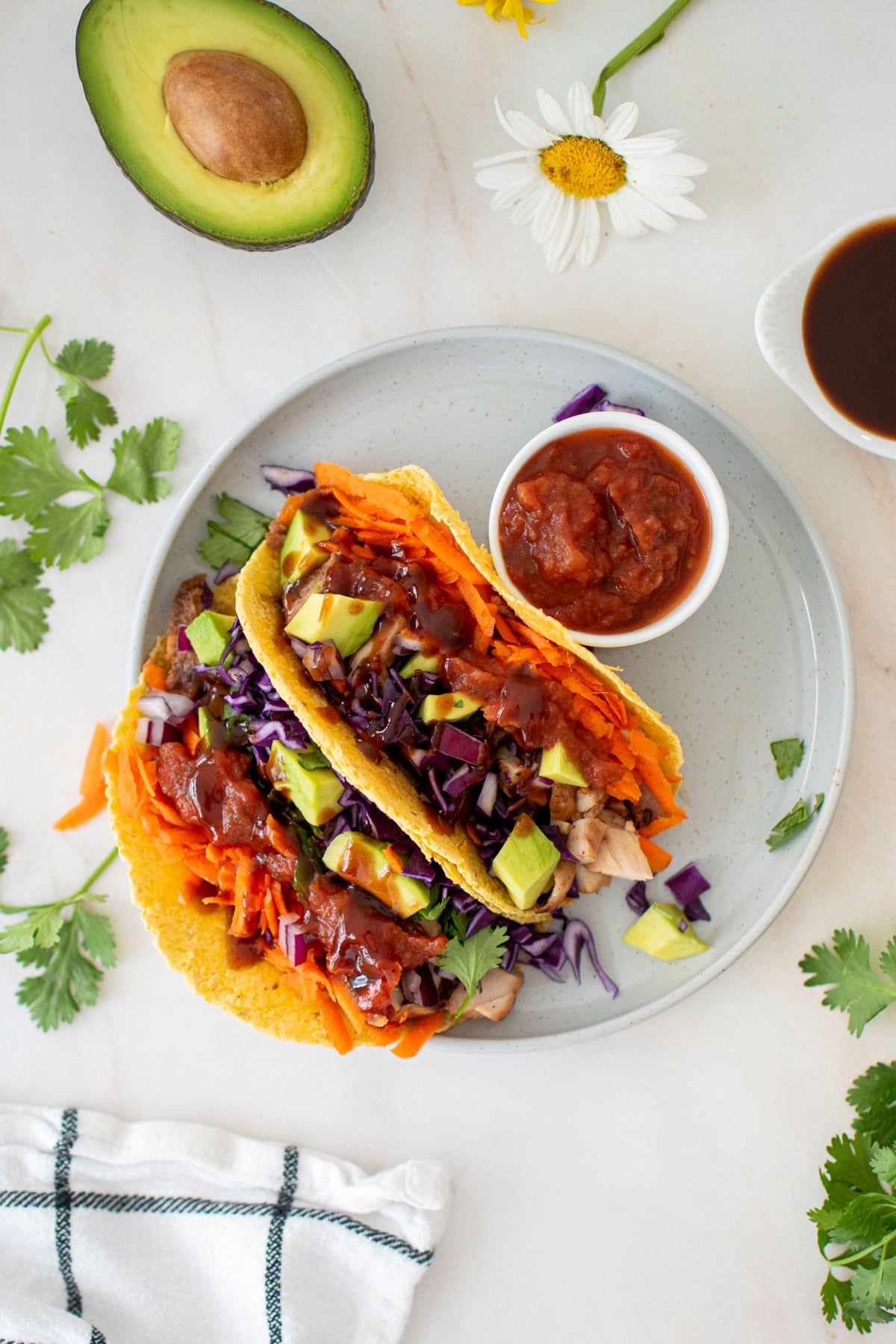 two bbq chicken tacos on a plate with a small dish of salsa on a marble counter with half an avocado and fresh flowers.