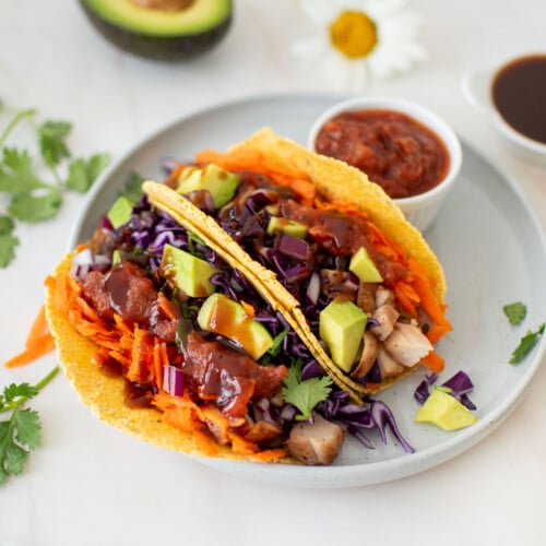 two bbq chicken tacos on a plate with a small dish of salsa on a marble counter with half an avocado and fresh flowers.