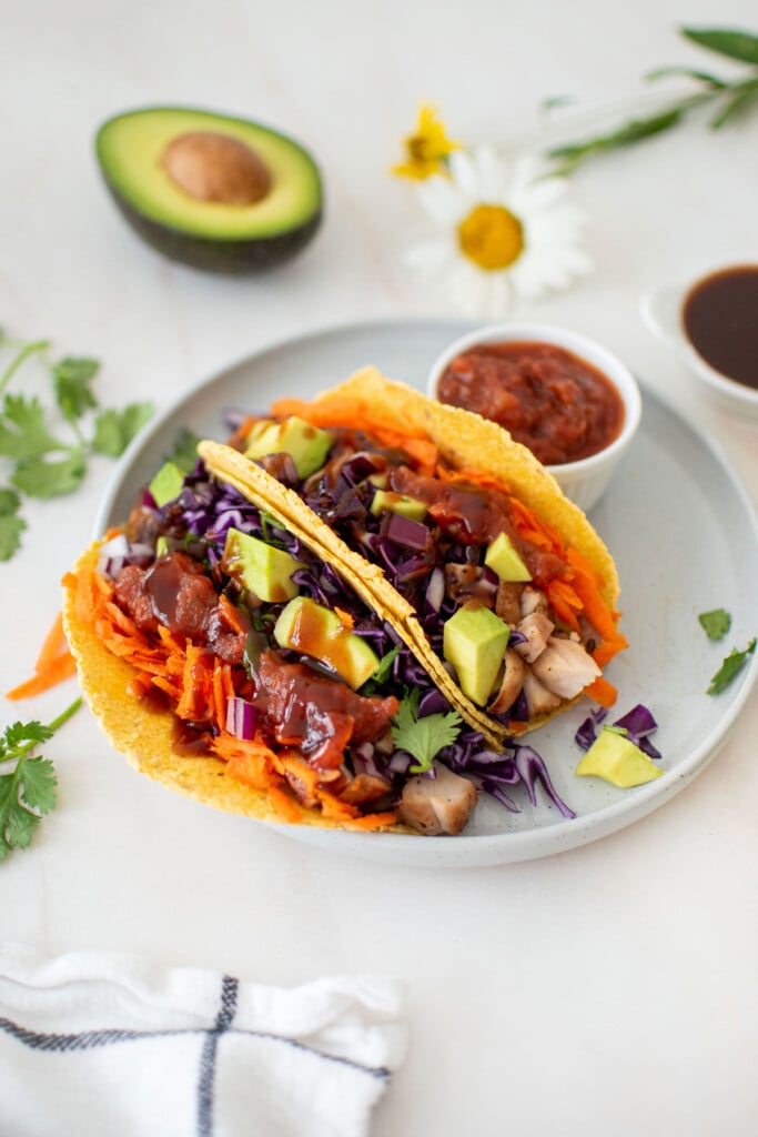 two bbq chicken tacos on a plate with a small dish of salsa on a marble counter with half an avocado and fresh flowers.