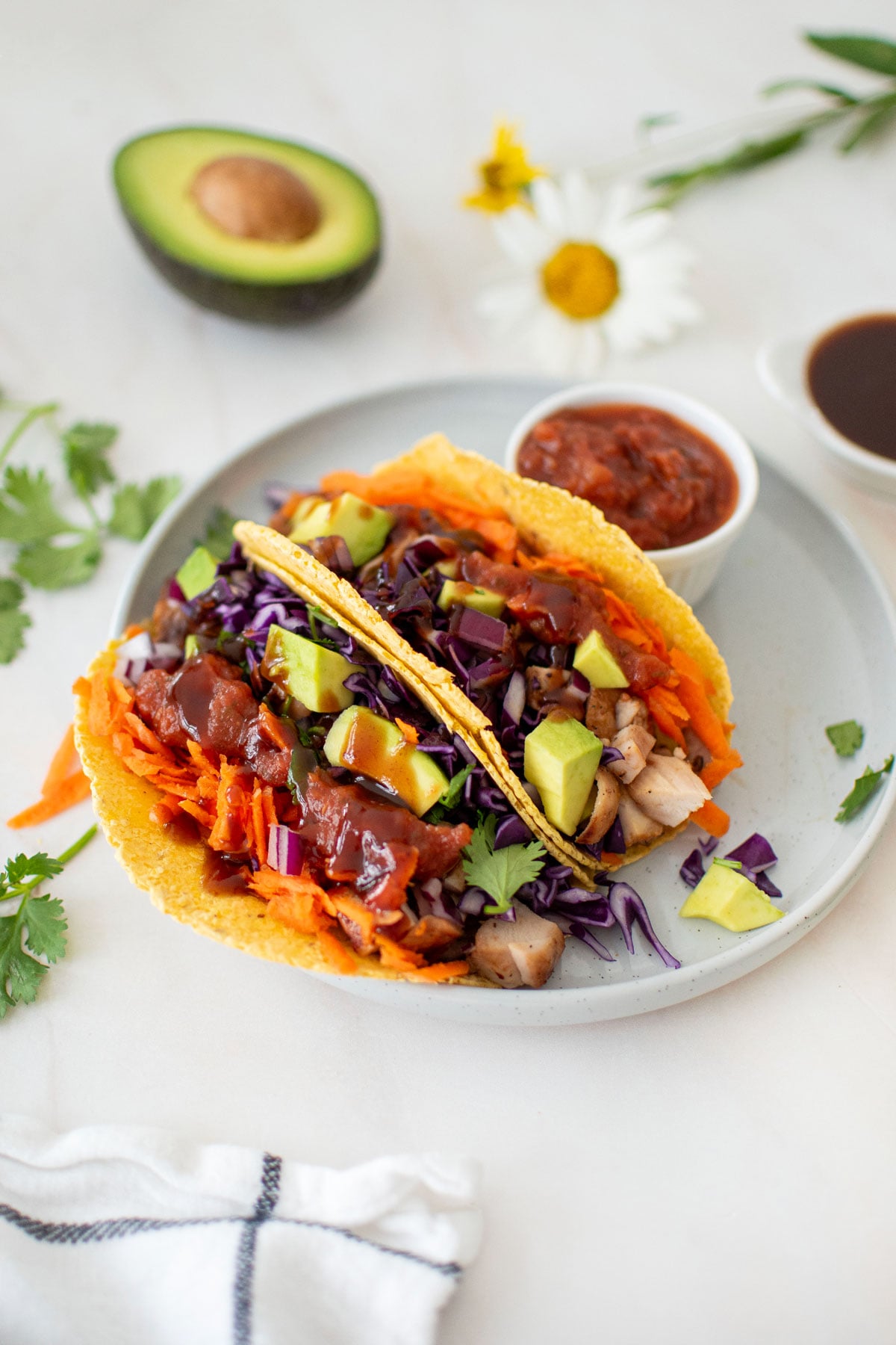 two bbq chicken tacos on a plate with a small dish of salsa on a marble counter with half an avocado and fresh flowers.