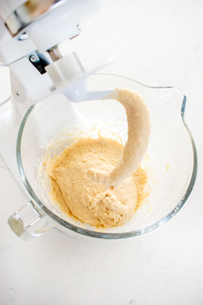 bagel dough mixed up in a glass mixing bowl on a white marble countertop.