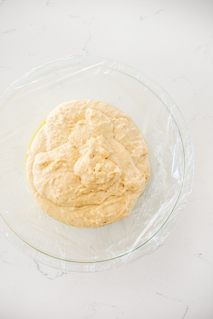bagel dough in a large glass bowl covered with plastic wrap on a white marble countertop.