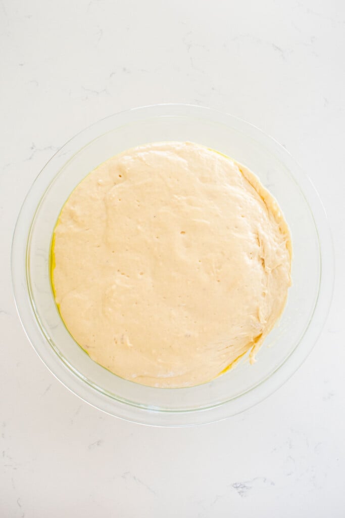 bagel dough that has risen in a large glass bowl on a white marble countertop.