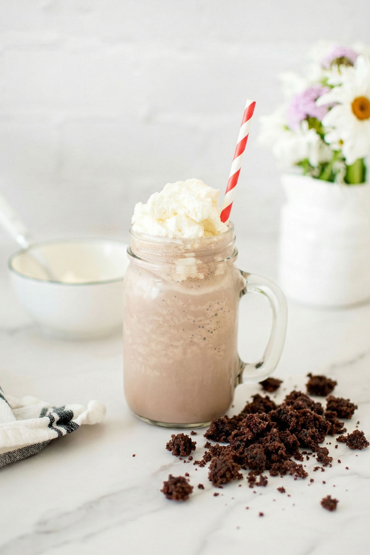 chocolate shake made with cake and fresh whipped cream and a straw on a white marble counter with flowers.