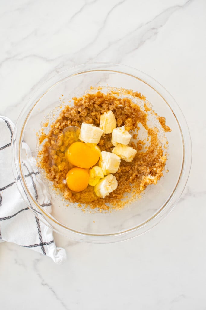creamed oil and sugars in a glass mixing bowl with banana chunks and eggs on a white marble counter.