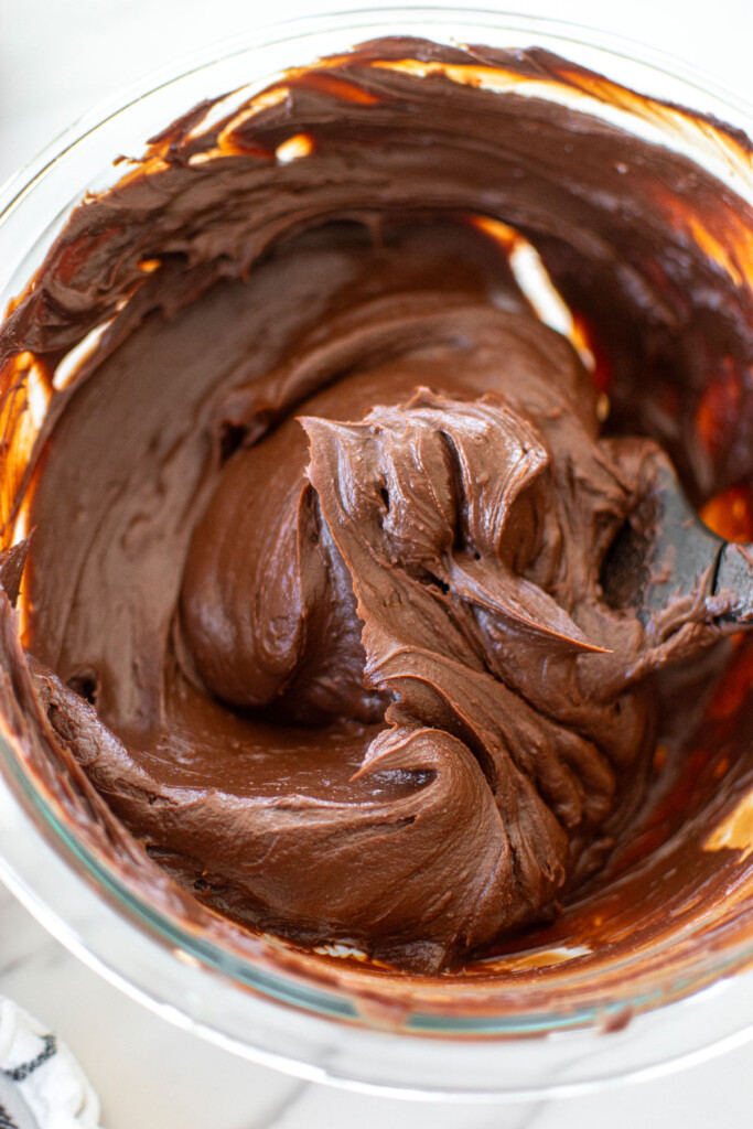 chocolate peanut butter caramel ganache frosting in a glass mixing bowl with a spatula on a white marble counter.