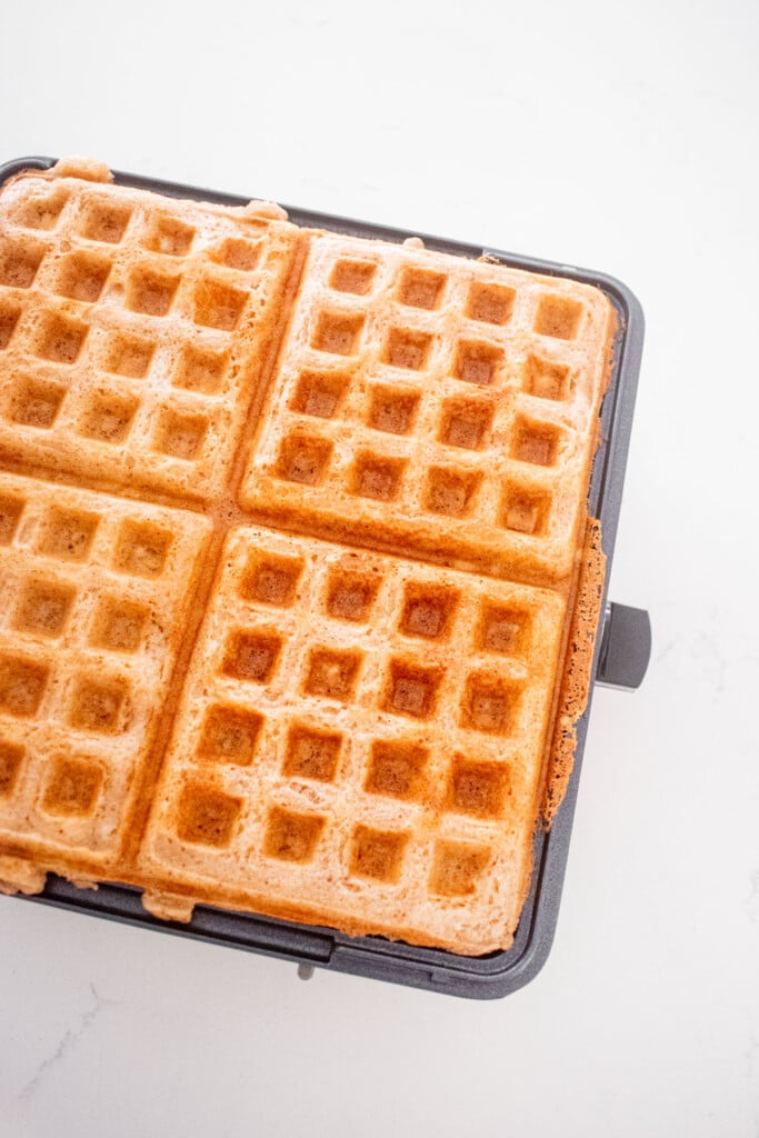cooked eggless apple cinnamon waffles in a waffle iron on a white marble counter.