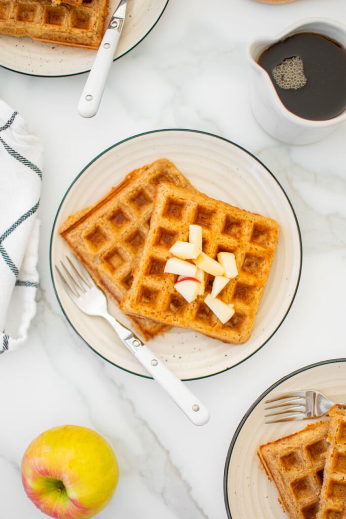 eggless apple cinnamon waffles on a plate with chopped apples on a white marble counter.