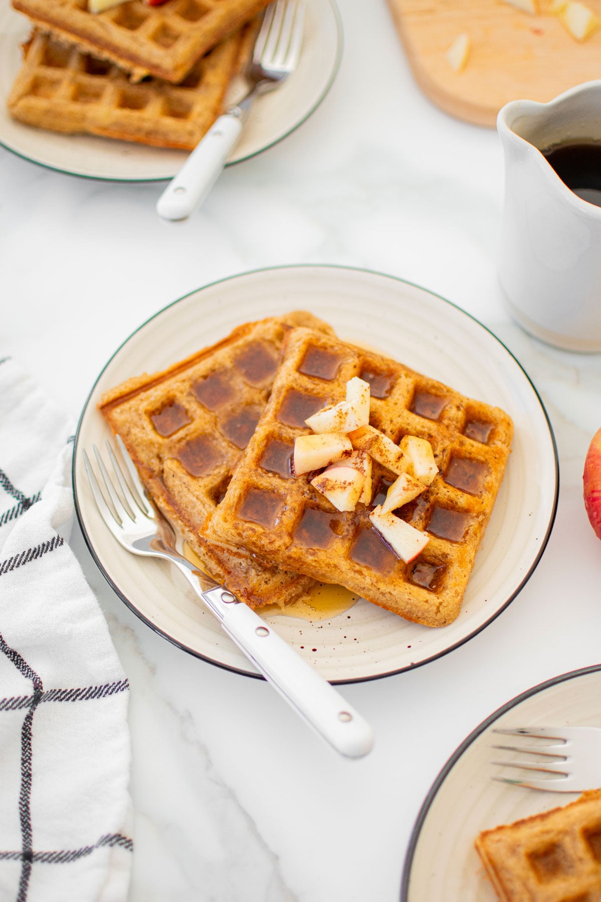 eggless apple cinnamon waffles on a plate with chopped apples and maple syrup on a white marble counter.