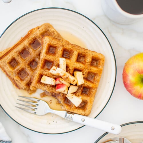 eggless apple cinnamon waffles on a plate with chopped apples and maple syrup on a white marble counter.