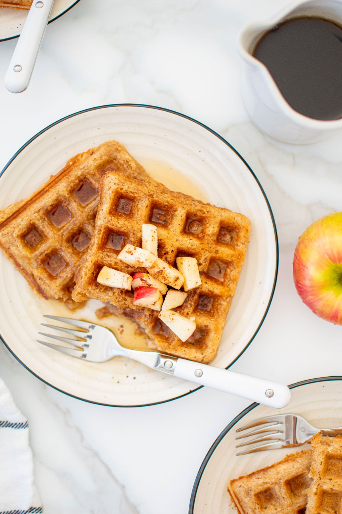 eggless apple cinnamon waffles on a plate with chopped apples and maple syrup on a white marble counter.