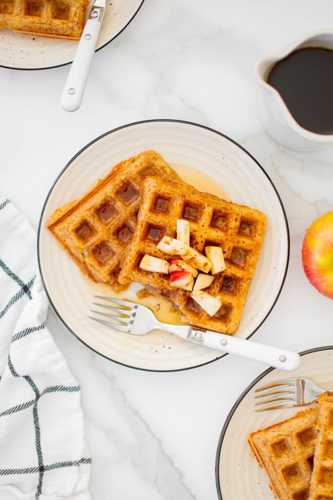 eggless apple cinnamon waffles on a plate with chopped apples and maple syrup on a white marble counter.