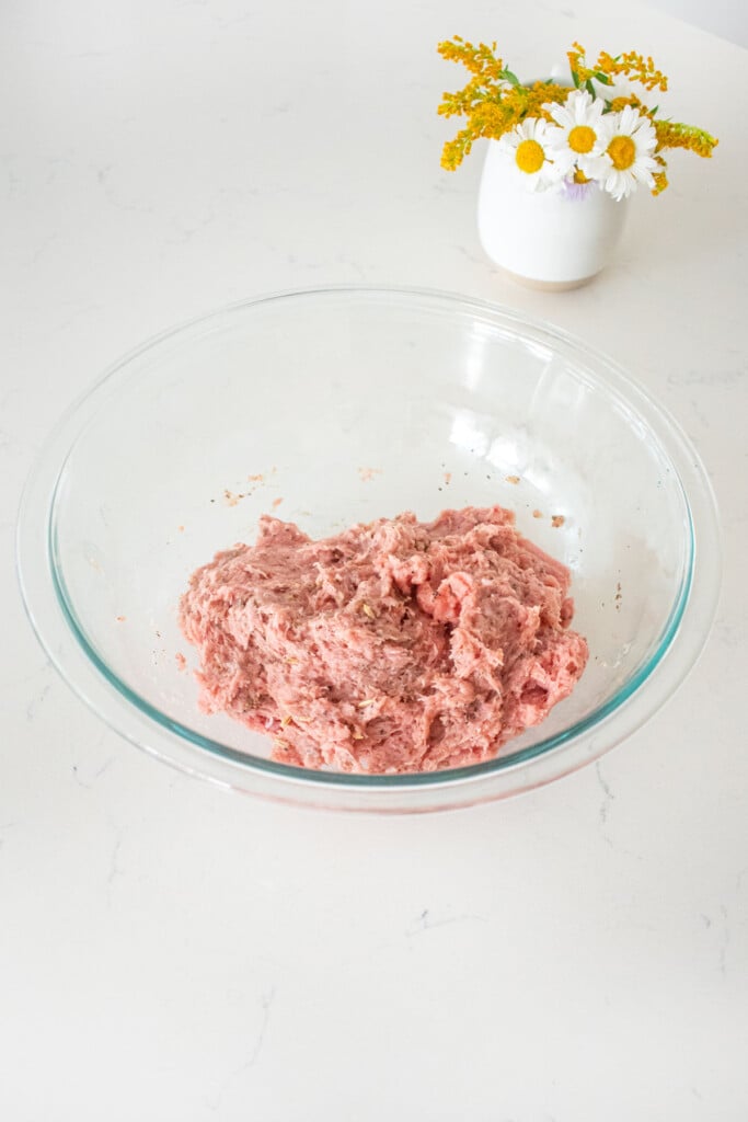 glass mixing bowl with ground turkey and spices mixed together on a marble counter with a small vase of fresh flowers.