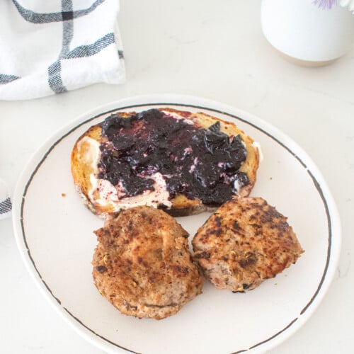 plate with two maple turkey breakfast sausage patties and a piece of sourdough toast with blueberry jam on a marble counter.