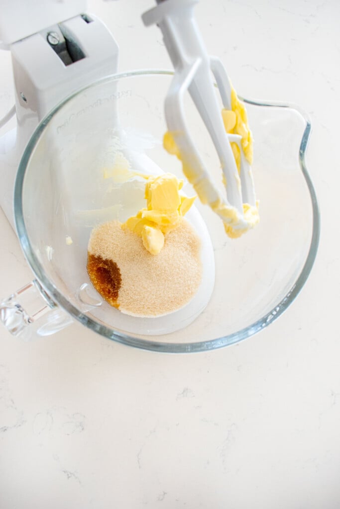 butter, cane sugar and vanilla extract in a glass stand mixing bowl on a white marble counter.