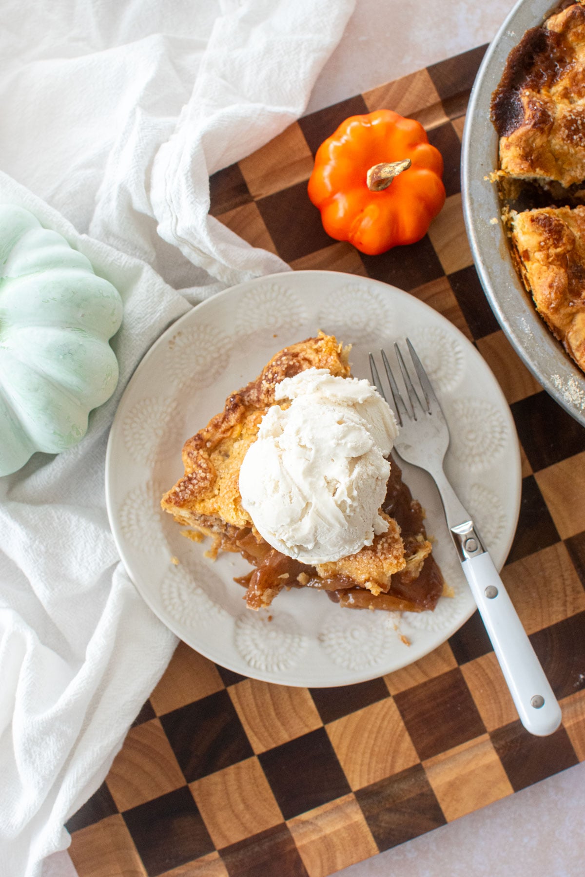 sourdough apple pie slice with sourdough discard pie curst and a scoop of vanilla ice cream on a white plate on a checkered wood cutting board.