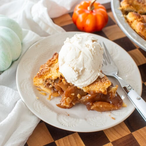 sourdough apple pie slice with a scoop of vanilla ice cream on a white plate on a checkered wood cutting board.