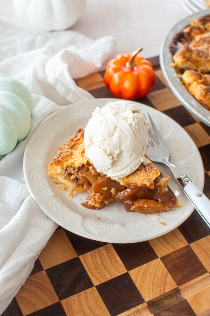 sourdough apple pie slice with a scoop of vanilla ice cream on a white plate on a checkered wood cutting board.