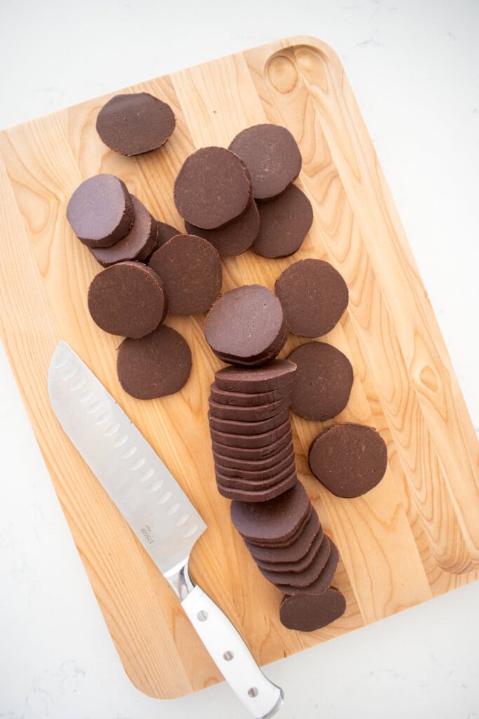 sourdough chocolate shortbread cookie dough sliced into cookie rounds on a wood cutting board on a white marble counter.