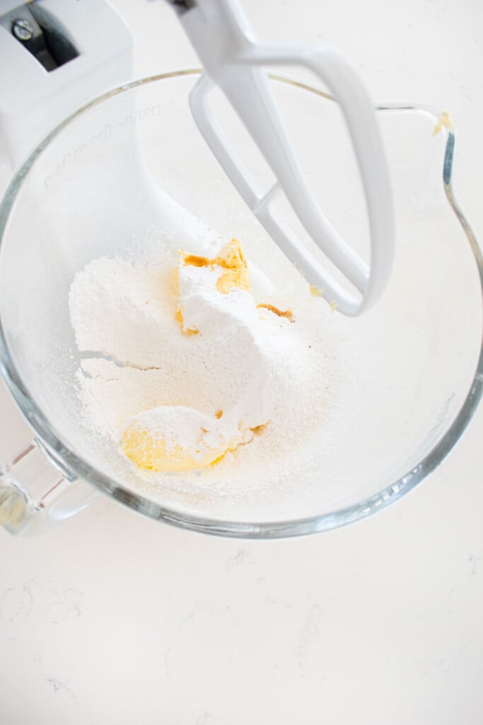 butter, powdered sugar and vanilla extract in a glass stand mixing bowl on a white marble counter.