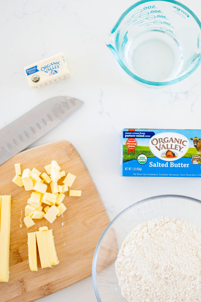 chopped butter on a wood cutting board with a bowl of flour on a white marble counter.