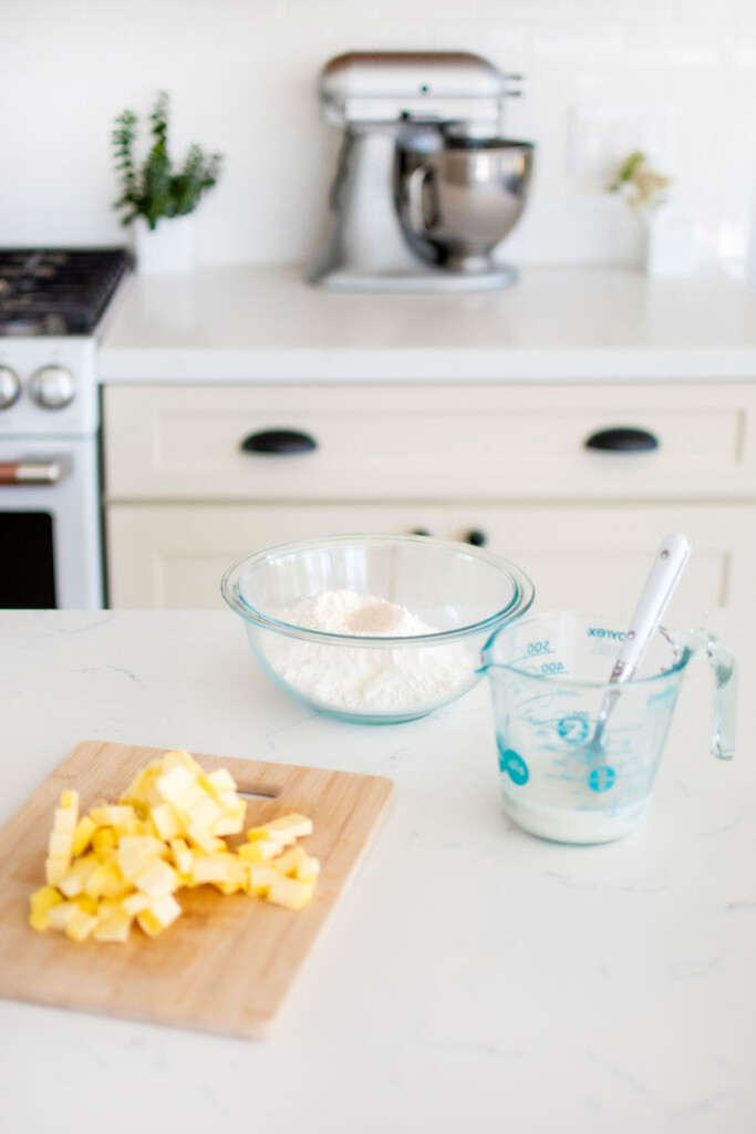 bowl of flour, wood cutting board with chopped butter, and a liquid measuring cup of sourdough discard and water on a white kitchen counter.