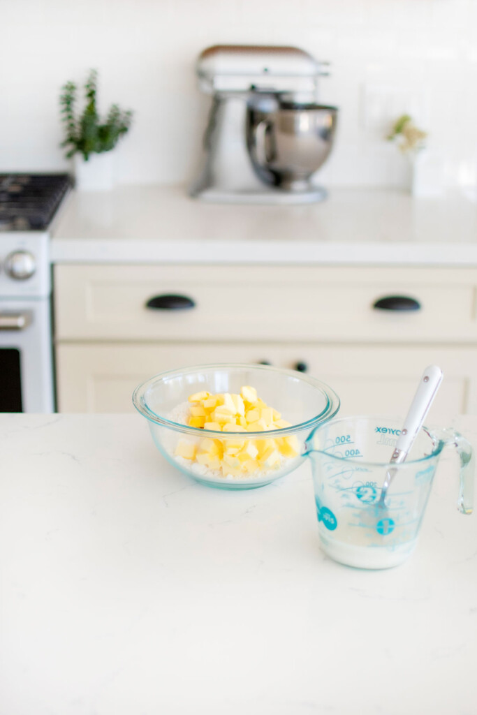 bowl of butter and flour with a liquid measuring cup of sourdough discard and water on a white kitchen counter.