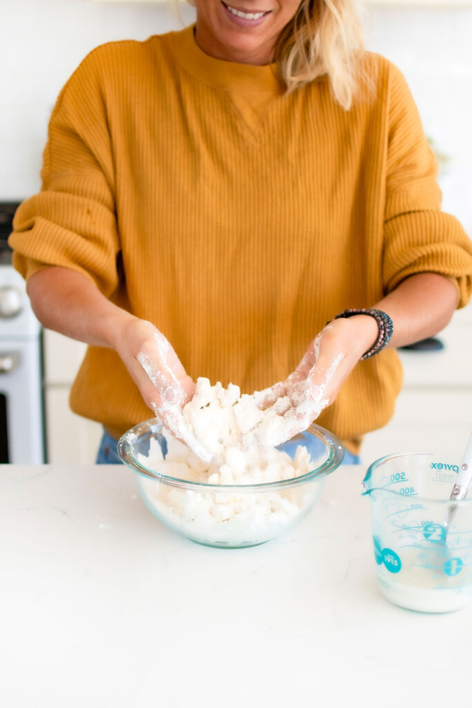 woman mixing sourdough pie crust in a glass mixing bowl with her hands at a white kitchen counter.