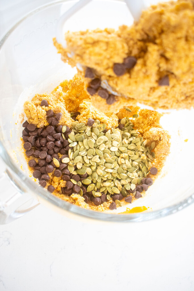 chocolate chips and pumpkin seeds in gluten free pumpkin cookie dough in a glass mixing bowl on a white marble counter.