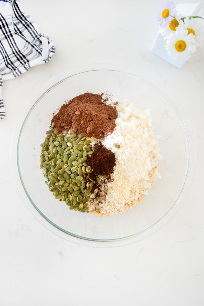 dry ingredients for chocolate olive oil granola in a glass mixing bowl on a marble counter with a small vase of flowers.