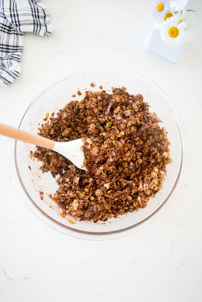 glass mixing bowl with chocolate olive oil granola mixed together on a marble counter.