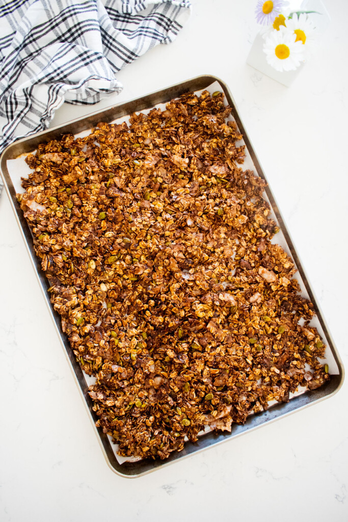 chocolate olive oil granola spread out onto a baking sheet on a marble counter with a small vase of flowers.