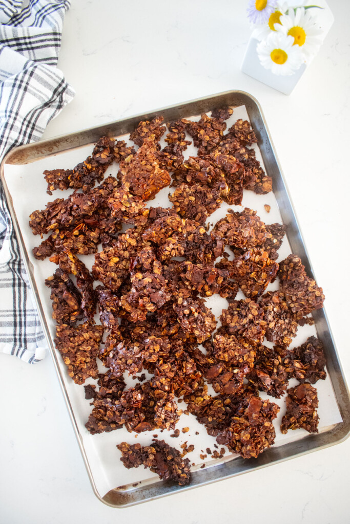 crispy chocolate olive oil granola clusters on a baking sheet on a marble counter with a small vase of flowers.