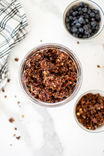 glass container of chocolate olive oil granola on a white marble counter.