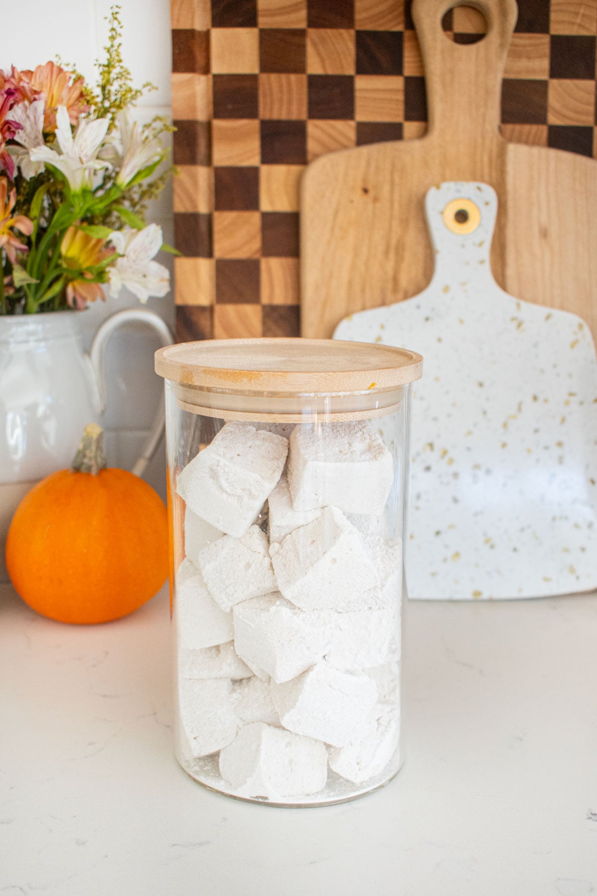 glass jar with homemade mini marshmallows inside sitting on a marble counter with fresh flowers and min pumpkins.