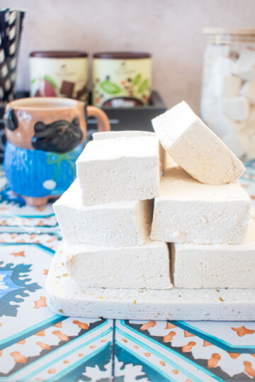 stack of homemade marshmallows on a marble serving board on a tile table with a mug of cocoa.