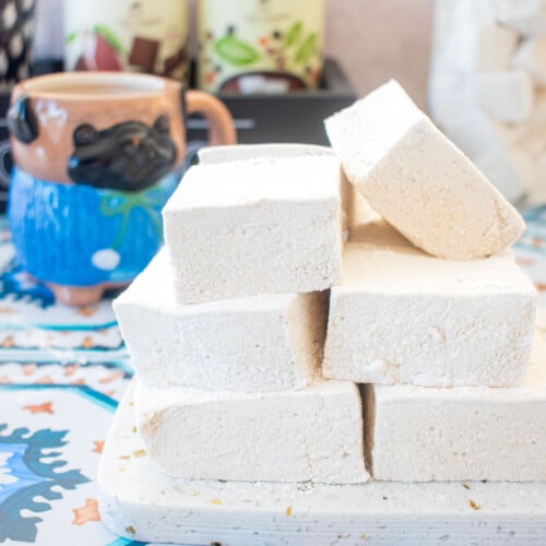 stack of homemade marshmallows on a marble serving board on a tile table with a mug of cocoa.