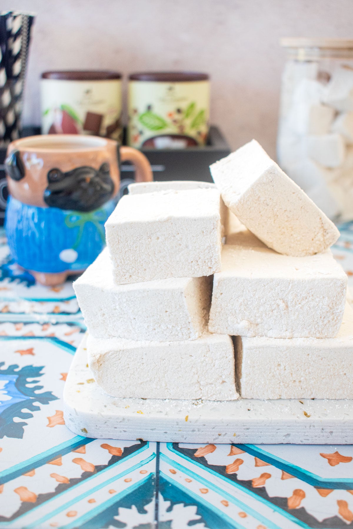 stack of homemade marshmallows on a marble serving board on a tile table with a mug of cocoa.