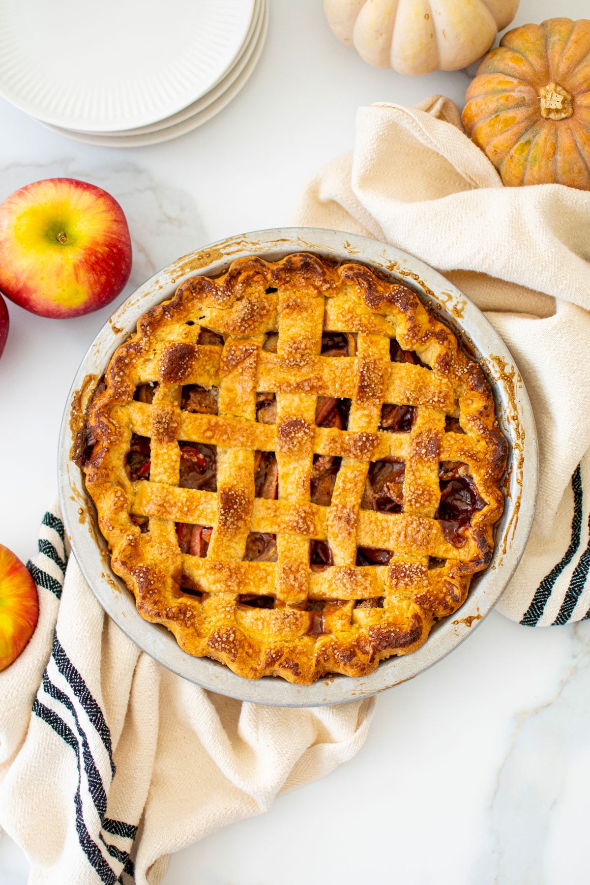 lattice spiced apple pie in a silver pie pan cut into slices on a white marble counter with apples and pumpkins.