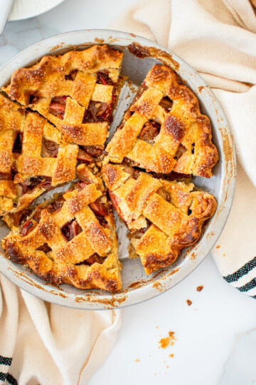 lattice spiced apple pie in a silver pie pan cut into slices on a white marble counter.