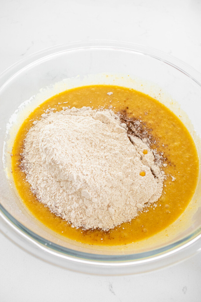 ingredients for pumpkin spice donut batter in a glass mixing bowl on a white marble counter.