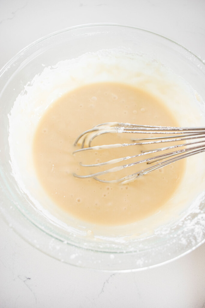 maple glaze in a glass mixing bowl with a whisk on a white marble counter.