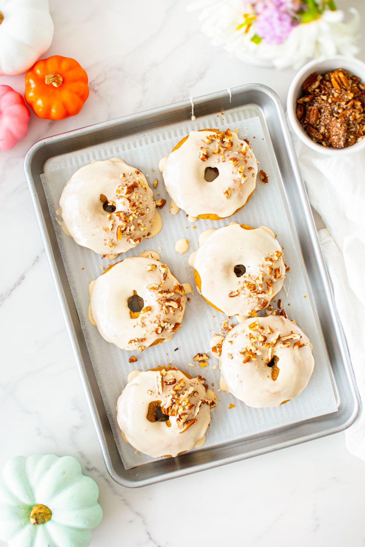 maple pumpkin spice donuts on a baking sheet on a white marble counter with fresh flowers and pumpkins.