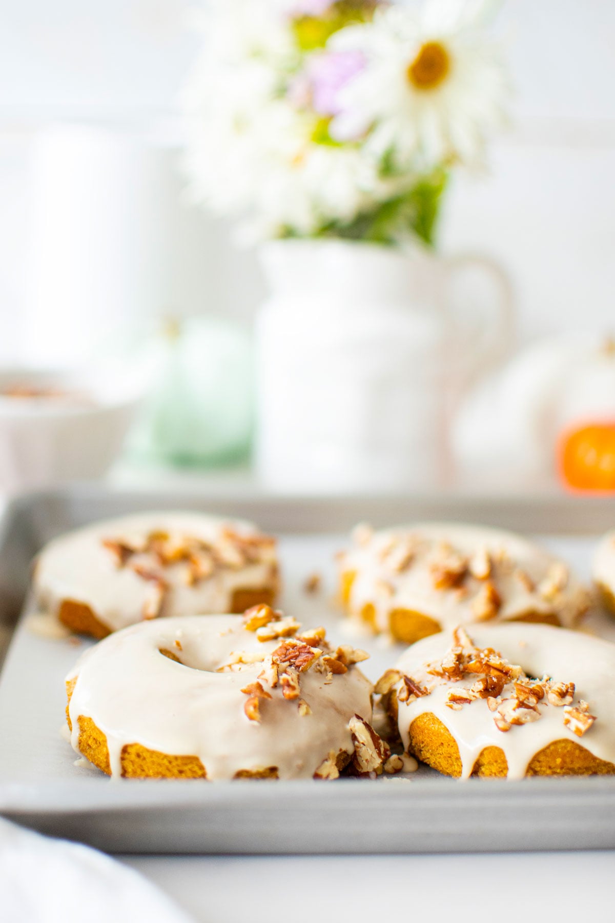 maple pumpkin spice donuts on a baking sheet on a white marble counter with fresh flowers and pumpkins.
