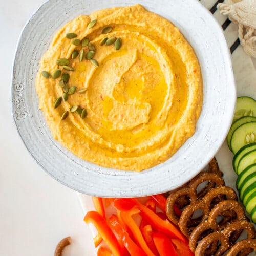 roasted red pepper hummus with purple garlic in a bowl on a charcuterie platter on a white marble table.