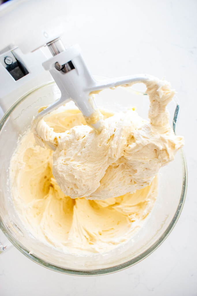 glass mixing bowl on a white marble counter with cream cheese frosting in the bowl and on the paddle.