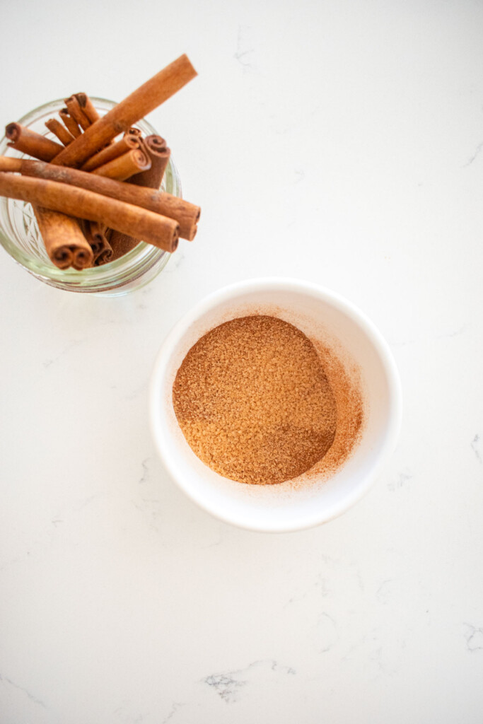 small bowl of cinnamon and sugar mixed together on a white marble counter with a little jar of cinnamon sticks.