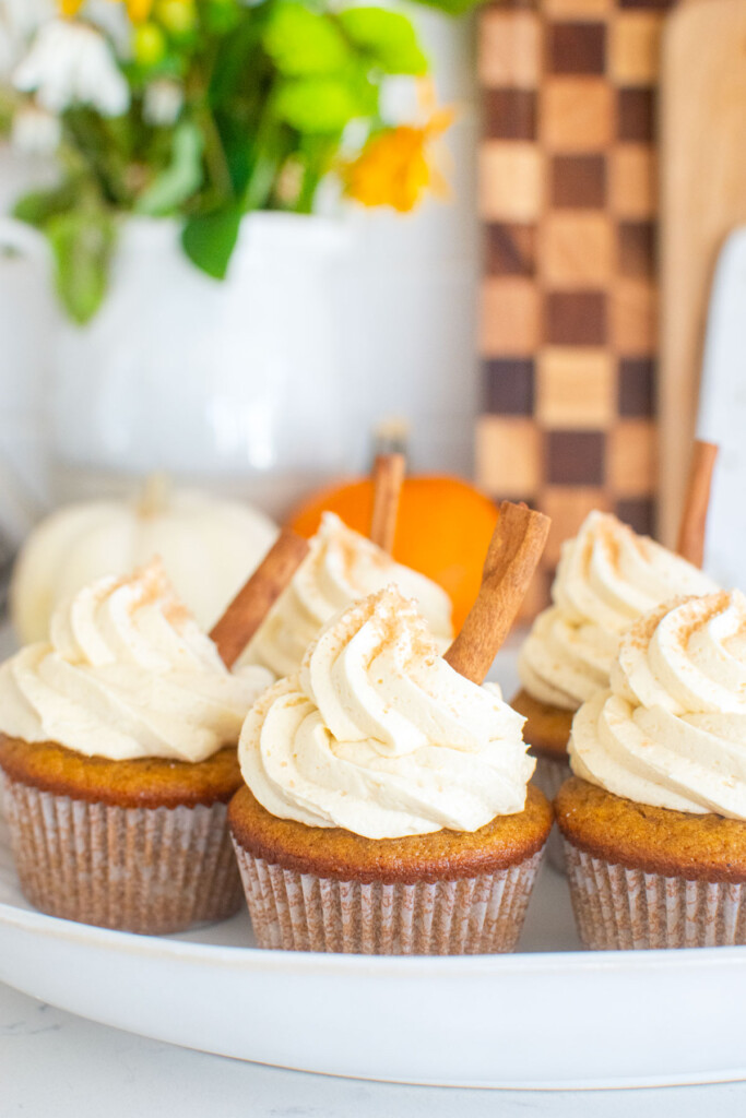 platter of sourdough pumpkin spice cupcake with cream cheese frosting and a cinnamon stick on a kitchen counter.