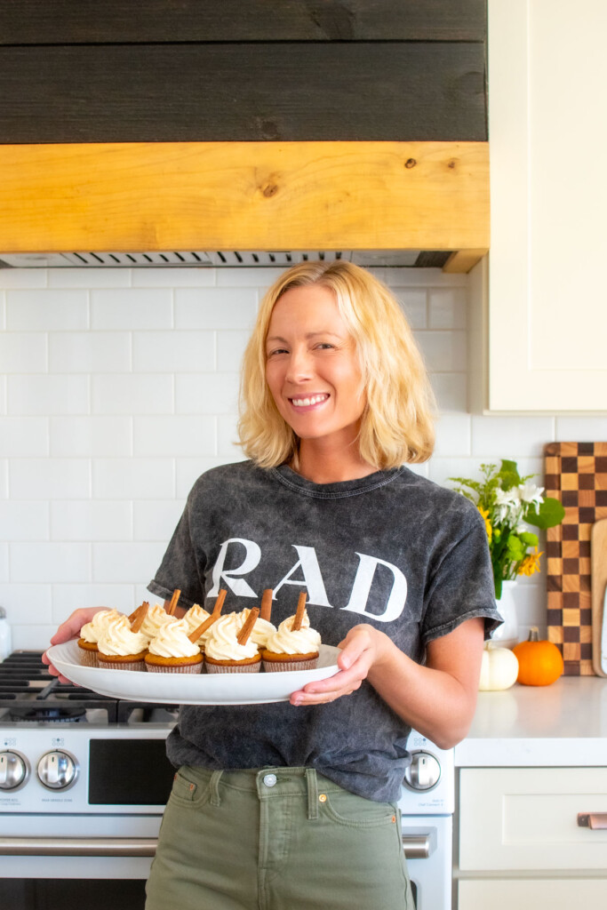 woman holding a platter of sourdough pumpkin spice cupcakes with cream cheese frosting and cinnamon sticks in the frosting in the kitchen.